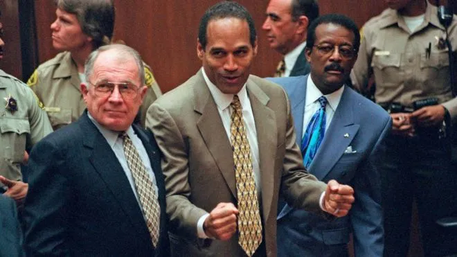 O.J. Simpson w garniturze, z zaciśniętymi pięściami, w sali sądowej w Los Angeles; obok niego obrońcy F. Lee Bailey i Johnnie Cochran, w tle policjanci