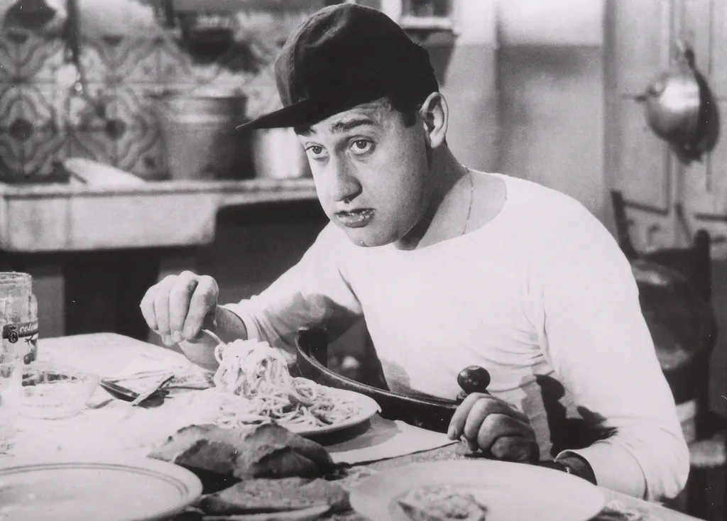 Czarno-białe ujęcie z filmu: Alberto Sordi w czapce z daszkiem nawija spaghetti na widelec i patrzy prosto w obiektyw