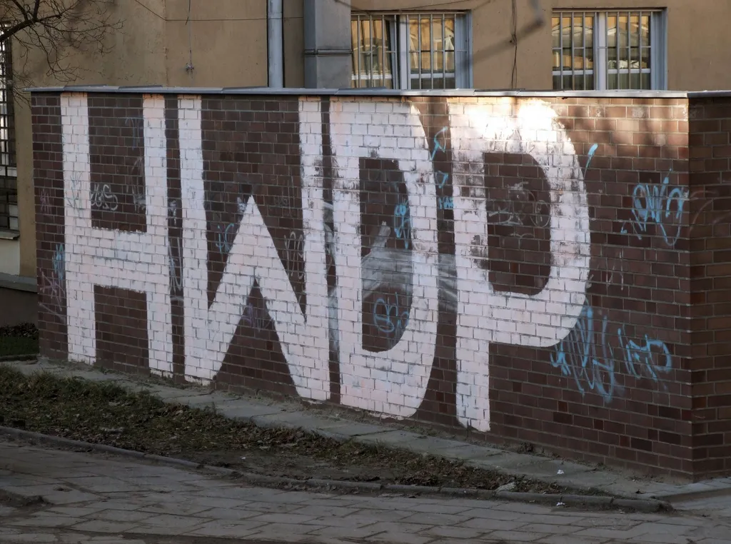 Wielki biały napis HWDP namalowany na ceglanym murze przy chodniku, wokół mniejsze niebieskie tagi graffiti