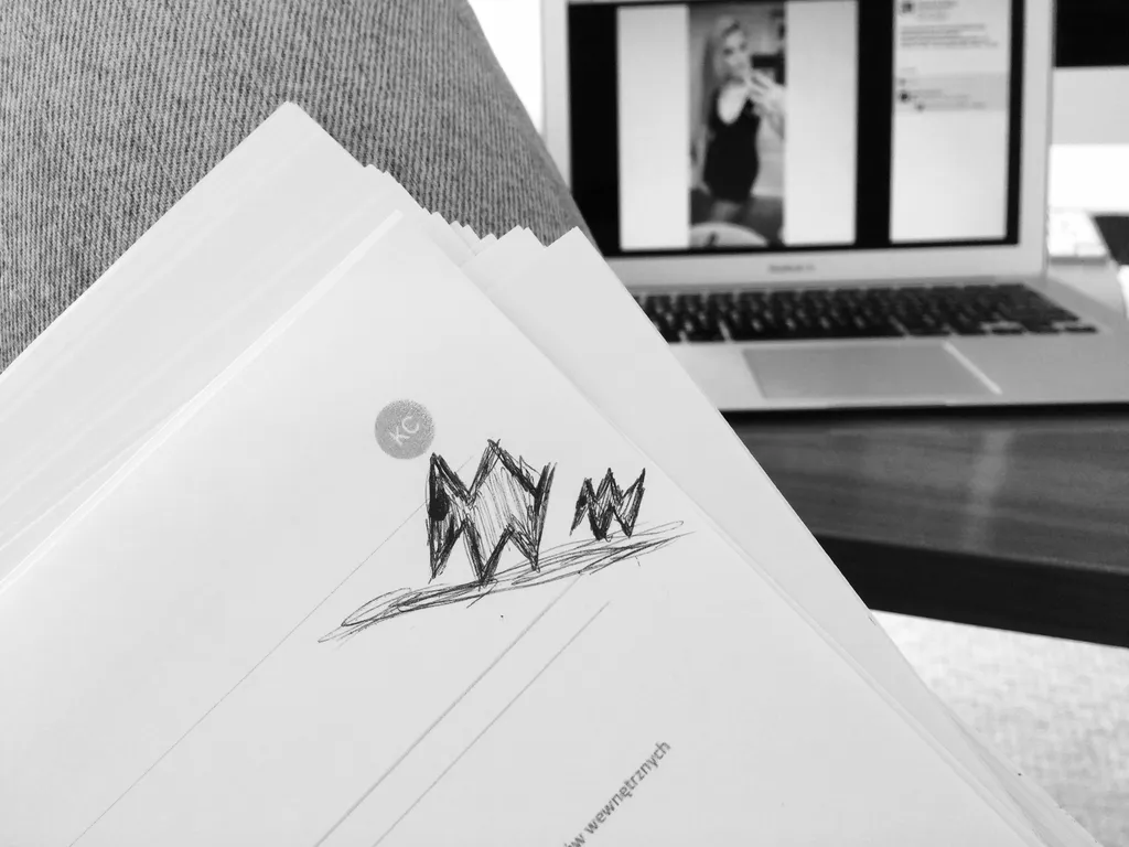 Plik wydrukowanych CV z rysunkiem długopisem na pierwszej stronie, w tle laptop z otwartym profilem kandydatki w serwisie społecznościowym