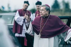 Janusz Gajos jako arcybiskup Mordowicz w purpurowej sutannie i z krzyżem na piersi gestykuluje przy samochodzie, za nim stoją dwaj duchowni