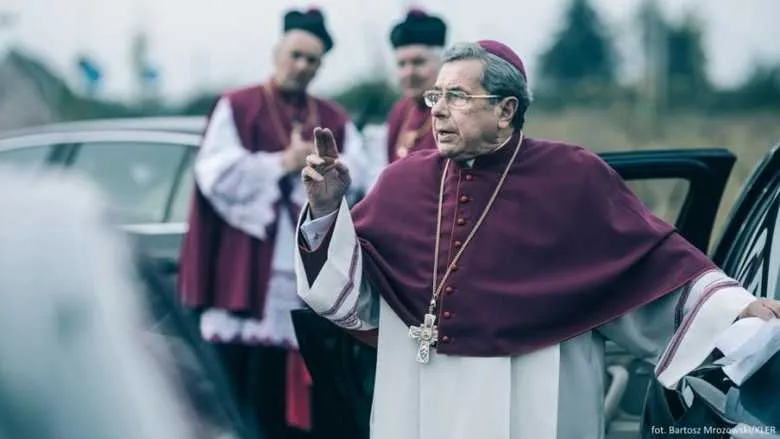 Janusz Gajos jako arcybiskup Mordowicz w purpurowej sutannie i z krzyżem na piersi gestykuluje przy samochodzie, za nim stoją dwaj duchowni