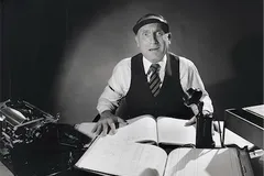 Czarno-białe zdjęcie zaskoczonego księgowego w daszku i kamizelce nad otwartymi księgami rachunkowymi, obok maszyna do pisania i telefon