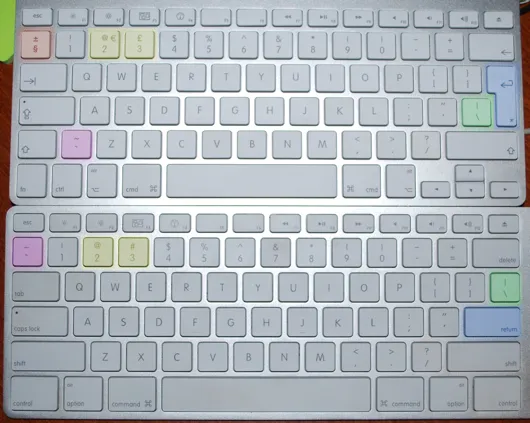 Dwie białe klawiatury Apple jedna nad drugą: u góry układ europejski ISO, na dole amerykański ANSI. Kolorami zaznaczono klawisze, które leżą w innych miejscach, m.in. Enter, tyldę i lewy Shift