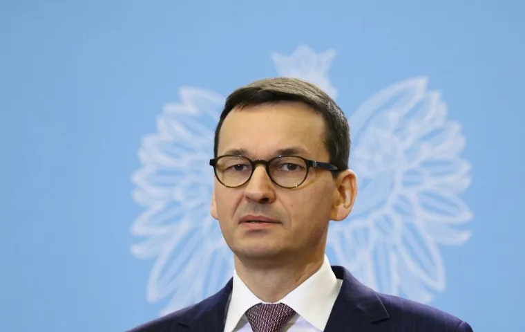 Mateusz Morawiecki w okularach, garniturze i krawacie na tle niebieskiej ścianki z białym orłem