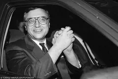 Czarno-białe zdjęcie uśmiechniętego Stana Tymińskiego w dużych okularach, siedzącego w samochodzie z uniesionymi, złączonymi dłońmi