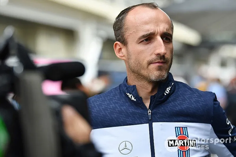 Robert Kubica w granatowo-białej bluzie zespołu Williams udziela wywiadu w padoku, przed nim kamera i mikrofon