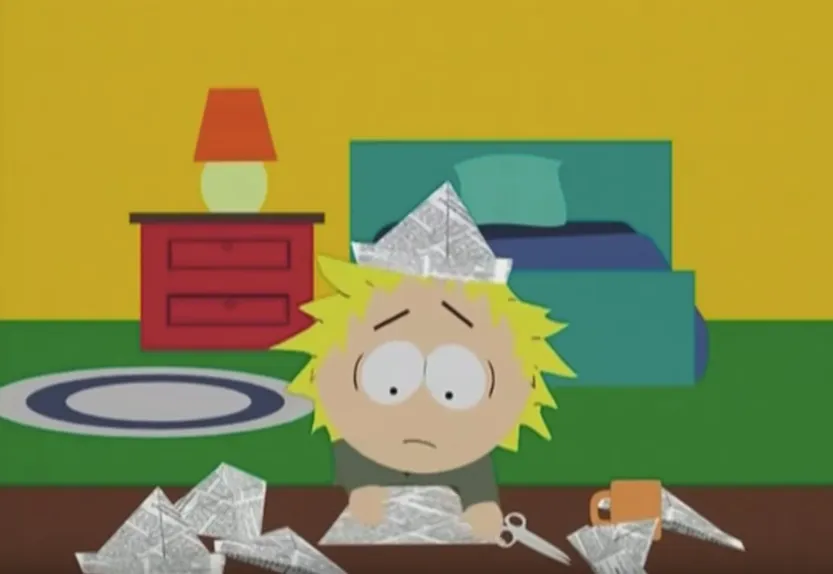 Zestresowany Tweek z serialu South Park siedzi przy biurku wśród papierowych łódek, z gazetową czapką na głowie